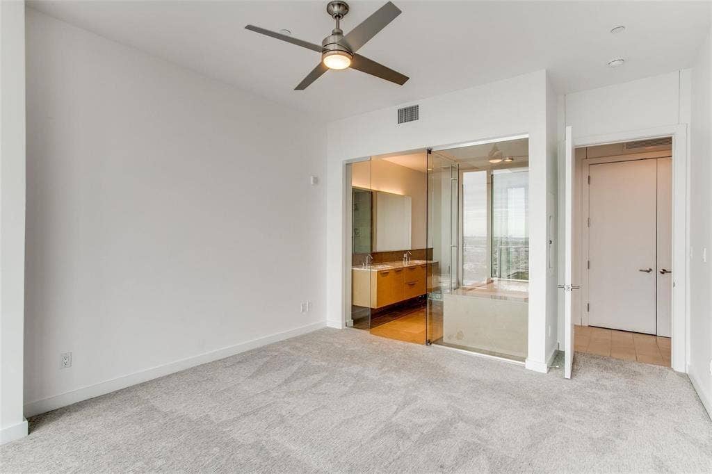 Spacious room with grey textured carpeting, contemporary ceiling fan, recessed lighting, and a full-height glass partition revealing a bathroom with a floating vanity and large soaking tub