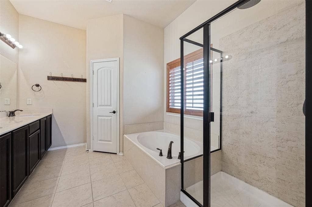 Dual vanity with dark wood-finish cabinetry, built-in soaking tub, walk-in shower with clear glass enclosure, and plantation shutters