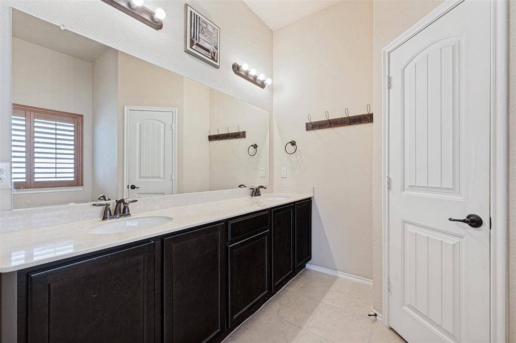 Dual undermount sinks with brushed bronze fixtures, dark wood-finish cabinetry, light-toned countertop, large wall-mounted mirror, and wood plantation shutters