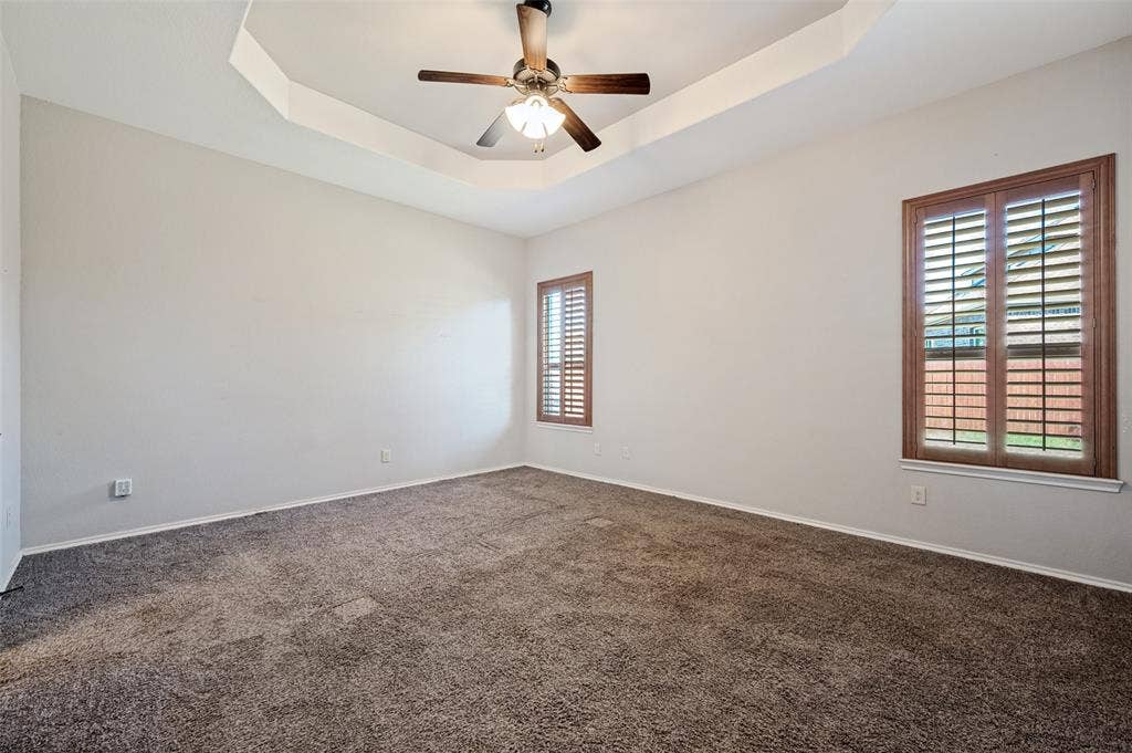 Tray ceiling with a ceiling fan light fixture, dual windows with wood-finish plantation shutters, and neutral-tone carpeting
