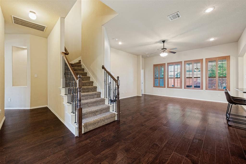 Hardwood-finish flooring throughout the main living area, staircase with carpeted treads and wrought iron balusters, four windows with wood shutters, recessed lighting, and a ceiling fan