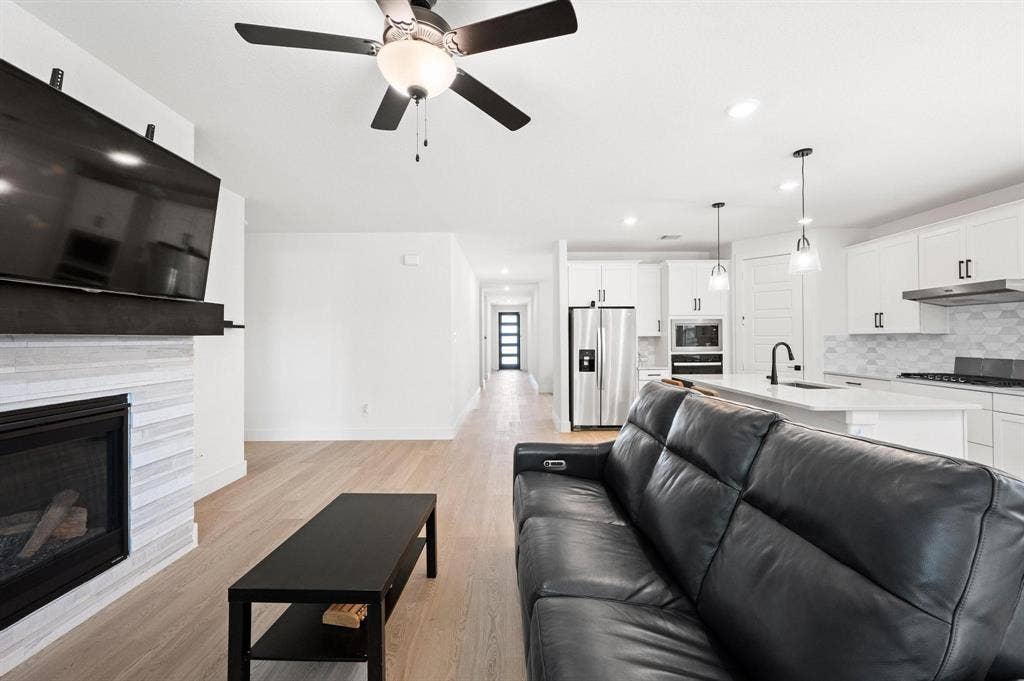 Integrated kitchen and living area featuring wood-finish flooring, a contemporary fireplace with tile surround, white cabinetry, stainless steel appliances, and an island with pendant lighting