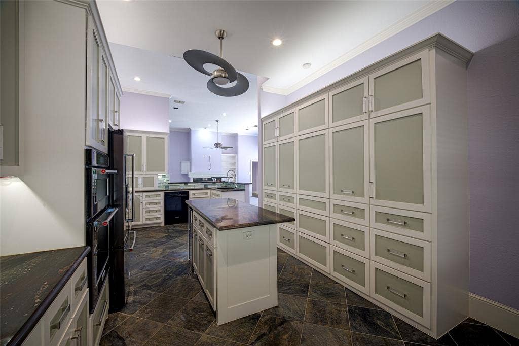 Full-height cabinetry with frosted glass inserts, dual wall-mounted ovens, a central island with countertop, and dark stone tile flooring