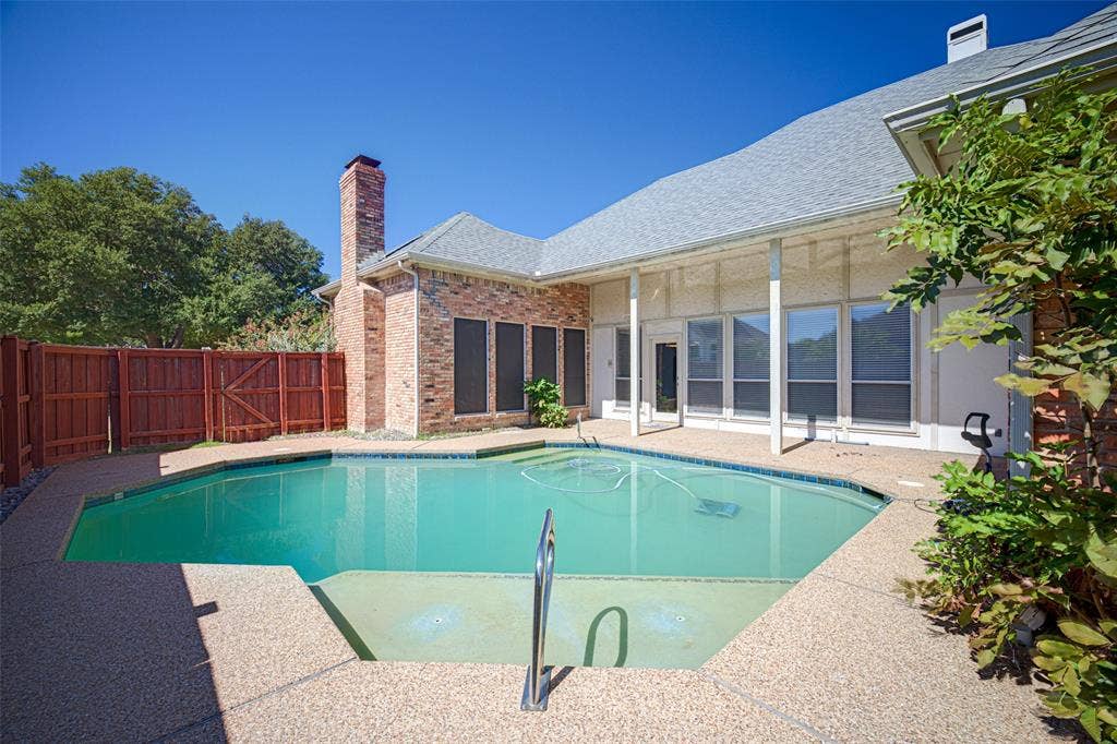 Sparkling resort-style pool with turquoise water and sun deck, framed by a brick chimney and expansive single-story residence featuring large glass sliding doors, a covered patio area, and mature landscaping with climbing vines along the perimeter