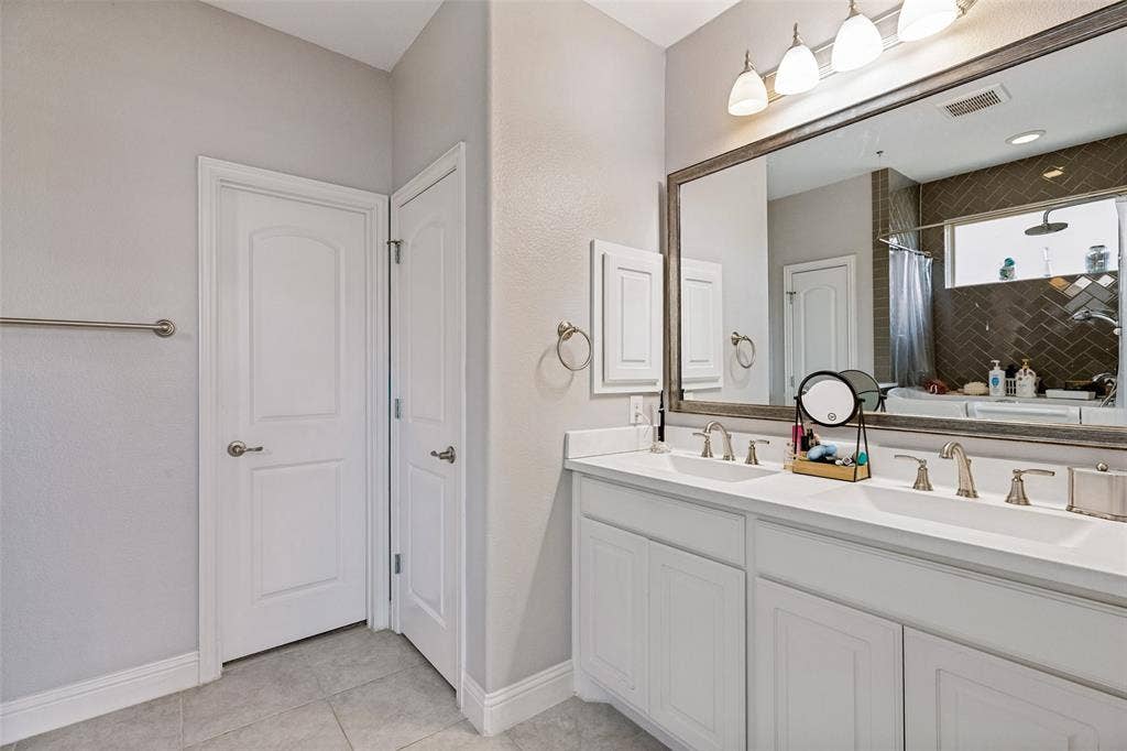 1004 Baynes Drive, McKinney, TX 75071 Dual vanity with white cabinetry and integrated sinks, a large framed mirror, and a tiled shower with a window