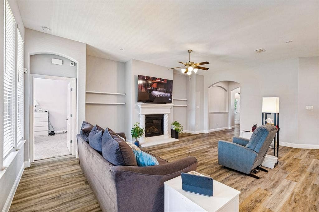 1004 Baynes Drive, McKinney, TX 75071 Spacious room featuring wood-finish flooring, a built-in fireplace with a white mantel, and ceiling fan with integrated lighting