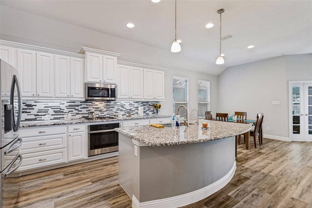 1004 Baynes Drive, McKinney, TX 75071 Open-concept kitchen featuring white cabinetry, stainless steel appliances, a large center island with a granite countertop, and wood-finish flooring