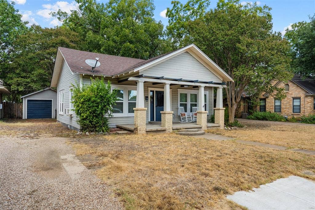 302 W Broadway Street, West, TX 76691 Gable roofline, covered front porch with stone-clad columns, horizontal siding, attached garage with dark blue door, and gravel driveway