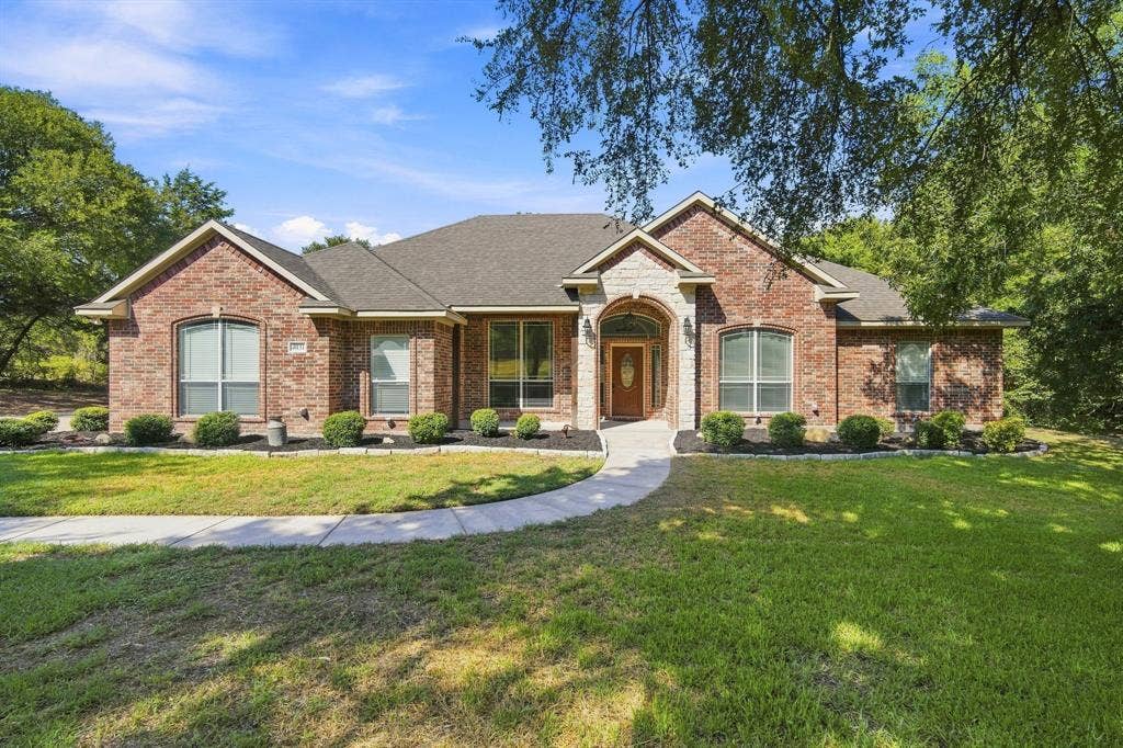 Stately brick home with a classic gabled roofline, framed by a lush, manicured lawn and mature shade trees.