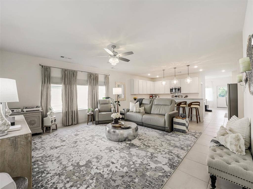Open-concept living area featuring a ceiling fan with light fixture, three large windows, a breakfast bar with pendant lighting, and ceramic tile flooring