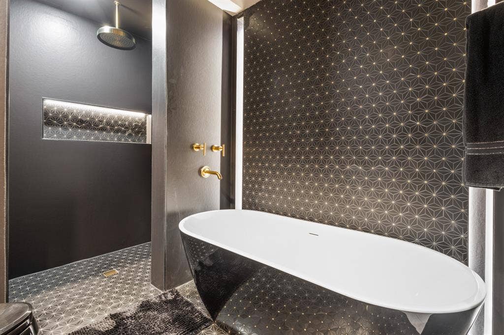 Spa-inspired primary bathroom featuring a freestanding oval soaker tub with integrated deck, geometric hexagonal tile work in warm bronze tones, rainfall showerhead, brass fixtures, and heated towel rack