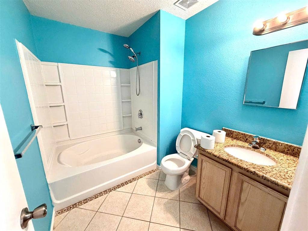 Tiled flooring with accent border, vanity with stone countertop and integrated sink, wall-mounted mirror, and a shower-tub combination with white surround