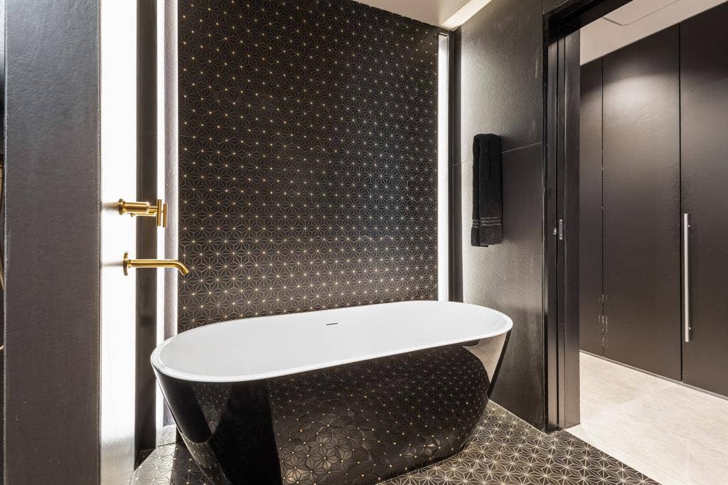2430 Victory Park Lane, #2401, Dallas, TX 75219 Freestanding black and white soaking tub, geometric pattern wall and floor tiles, gold-finish wall-mounted faucet, integrated vertical lighting, and built-in matte black cabinetry