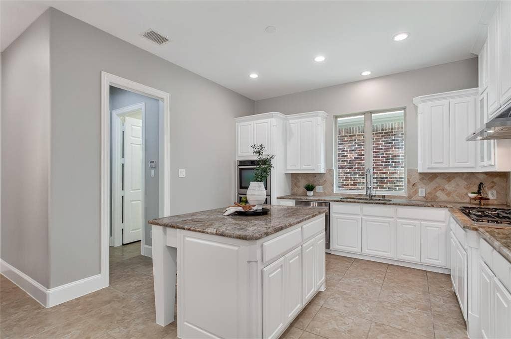 844 Veneto Drive, Allen, TX 75013 Undermount sink with a pull-down faucet set below a window, flanked by white raised-panel cabinetryand granite counters. Stainless dishwasher, double wall ovens, and a doorway to the utility hall.