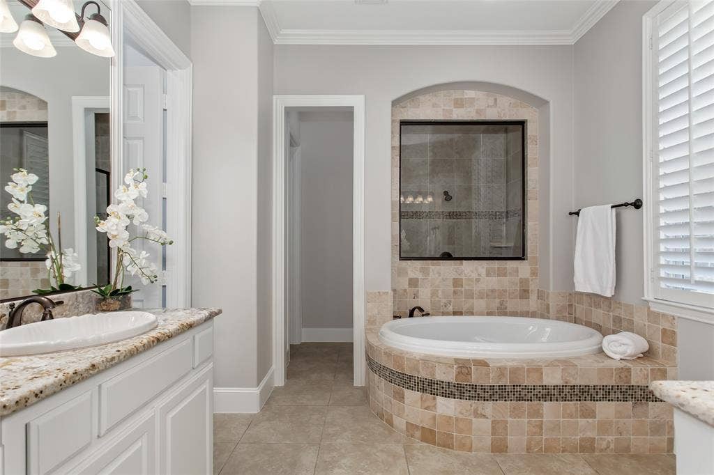 844 Veneto Drive, Allen, TX 75013 Garden tub with a tumbled travertine surround and glass mosaic accent band set below an archedniche with a glass window into the shower. Granite vanity, oil-rubbed bronze fixtures, and plantationshutters.