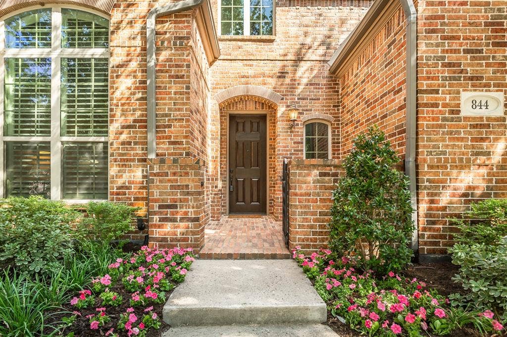 844 Veneto Drive, Allen, TX 75013 Arched brick entry with a wrought iron gate, brick-paver courtyard, and dark stained front door. Flowerbeds with fresh plantings from the 2026 landscape refresh frame the walkway.