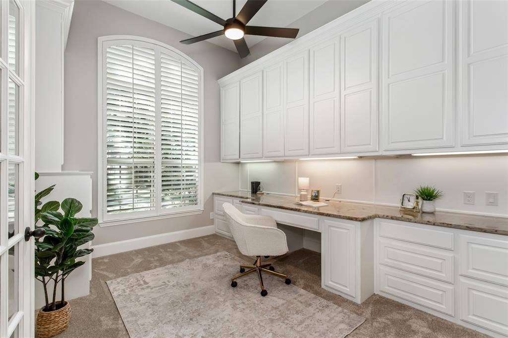 844 Veneto Drive, Allen, TX 75013 Private study with a wall of built-in cabinetry, a granite desktop with under-cabinet lighting, and anarched window with plantation shutters. New 2026 carpet and a new ceiling fan with an integrated LEDlight.