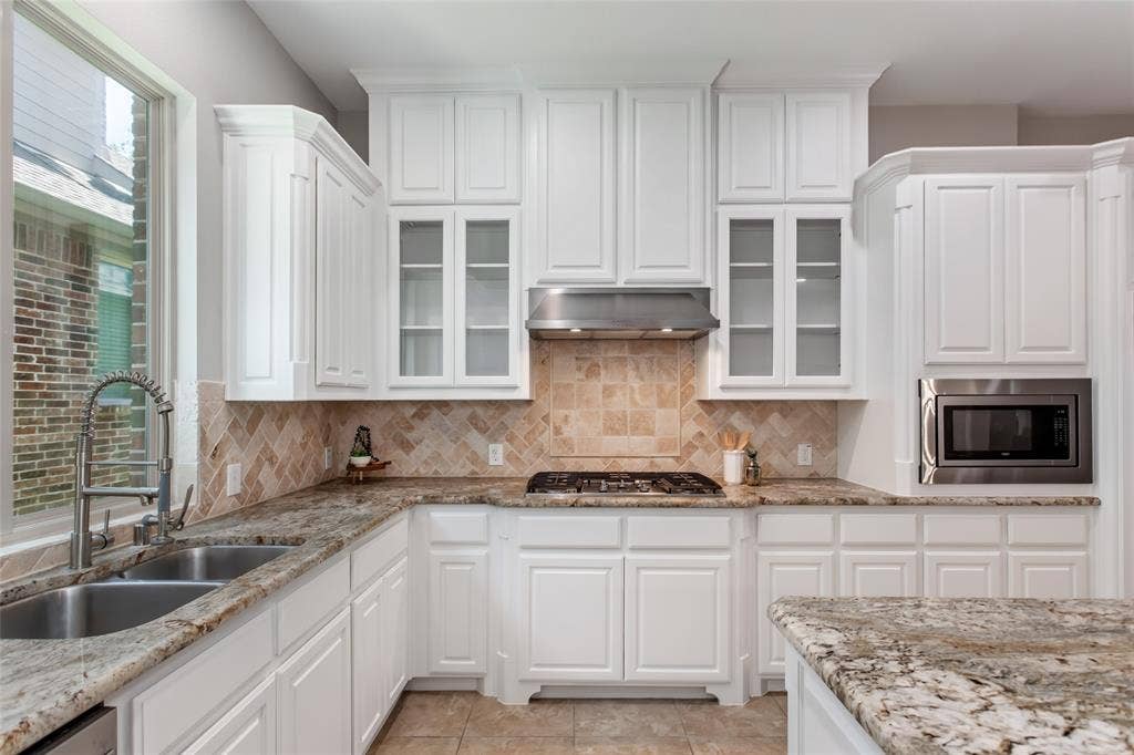 844 Veneto Drive, Allen, TX 75013 Gas cooktop with a stainless vent hood set against a tumbled stone backsplash with herringbone inlay.Glass-front upper cabinets, built-in microwave, walk-in pantry door, and island seating.