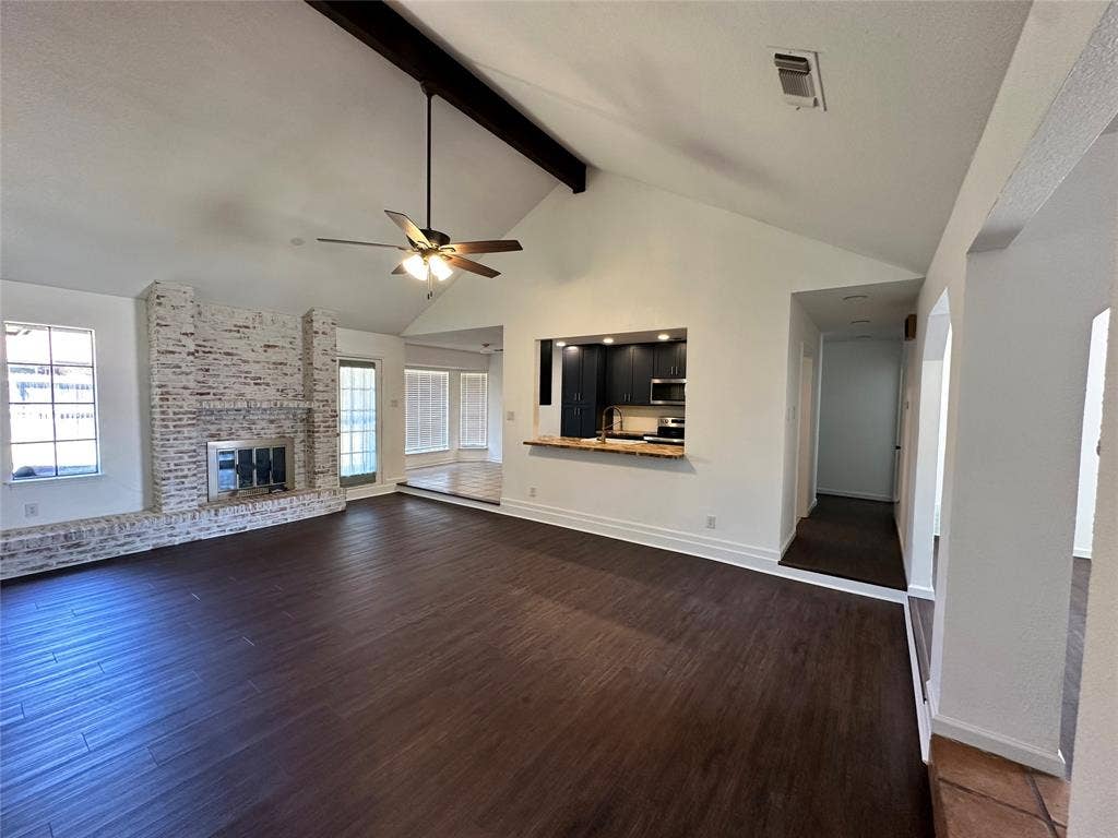 Vaulted ceiling with exposed beam, wood-finish flooring, white-painted brick fireplace, built-in kitchen pass-through, and ceiling fan