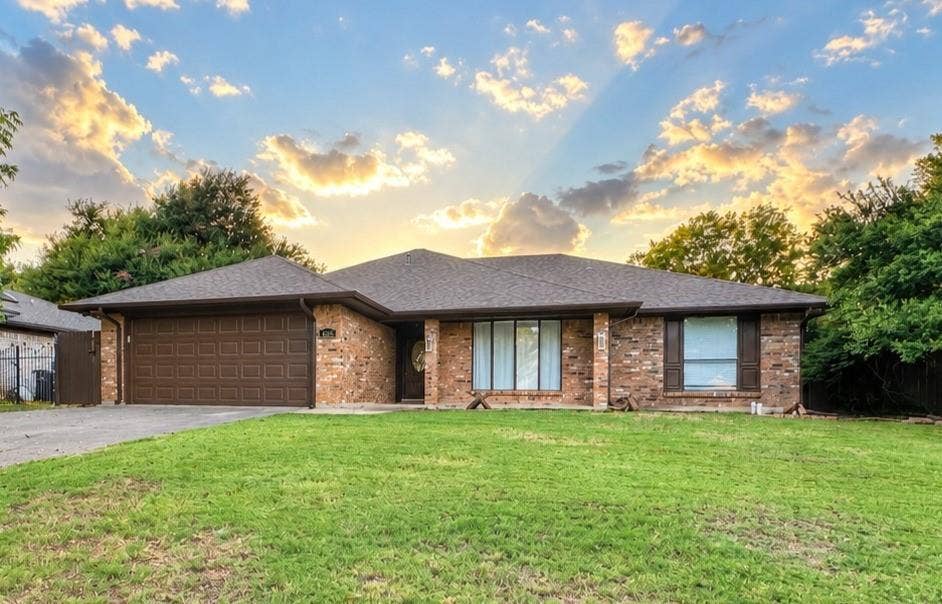 Brick exterior with a brown garage door, dark roof shingles, and mature front yard landscaping