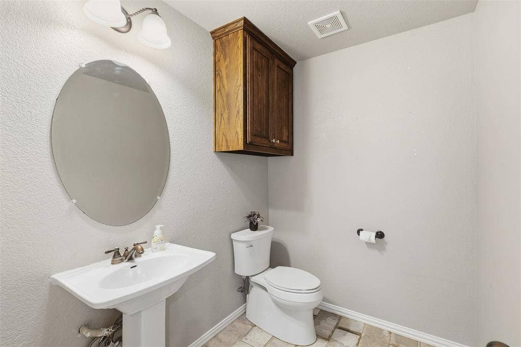 Pedestal sink with bronze-finish faucet, oval mirror, wall-mounted wood-finish cabinetry, and neutral-toned floor tiling