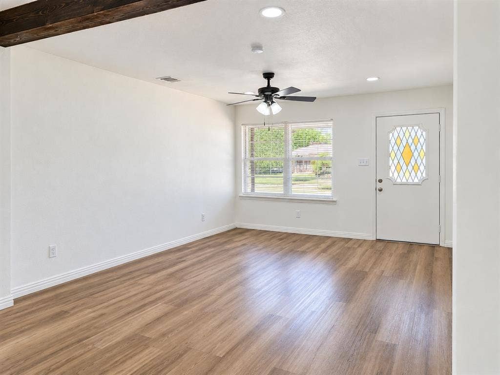 Wood-finish flooring, white walls, exposed wood beam, ceiling fan, and front entry door with leaded glass insert