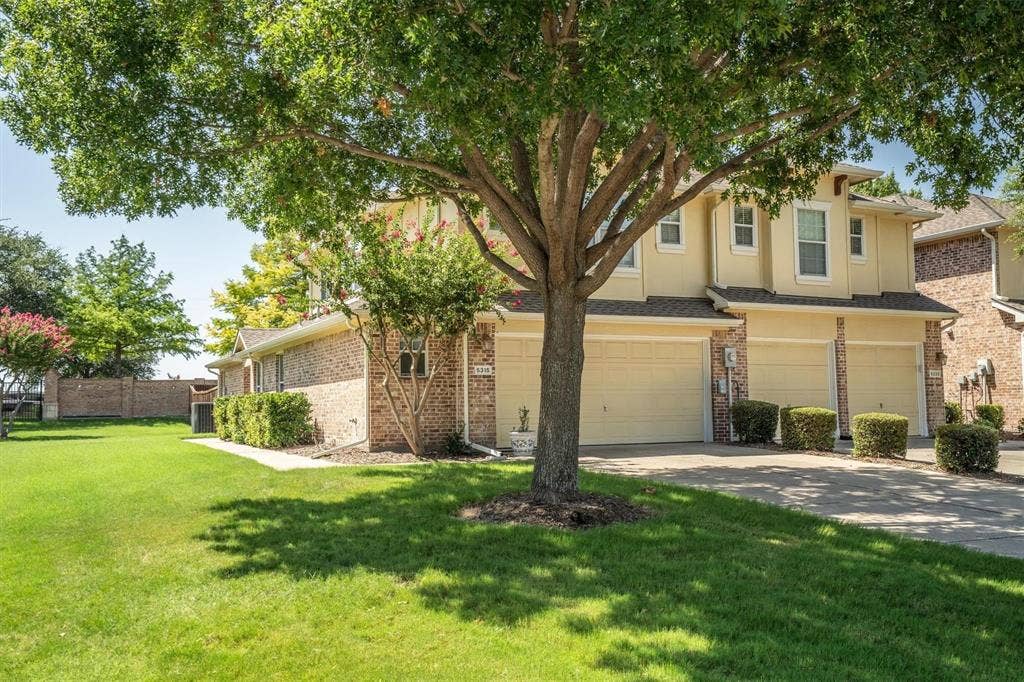 Brick and stucco exterior with multiple garage bays, featuring mature trees and landscaped shrubbery
