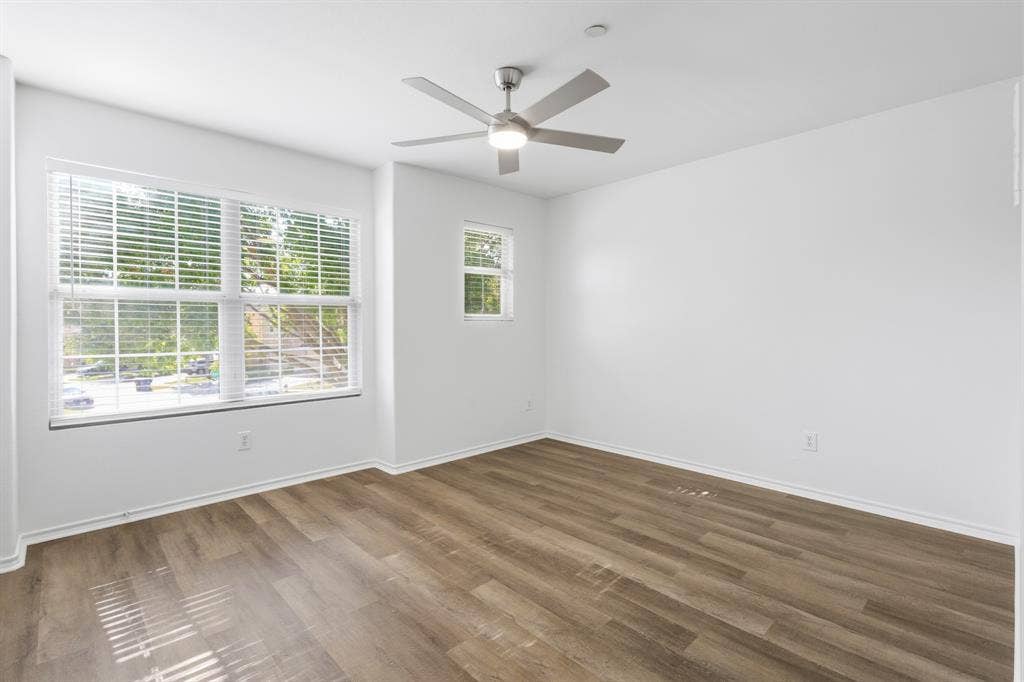 Spacious upstairs bedroom featuring wood-finish flooring, multiple windows with blinds, a modern ceiling fan with lighting, and white painted walls