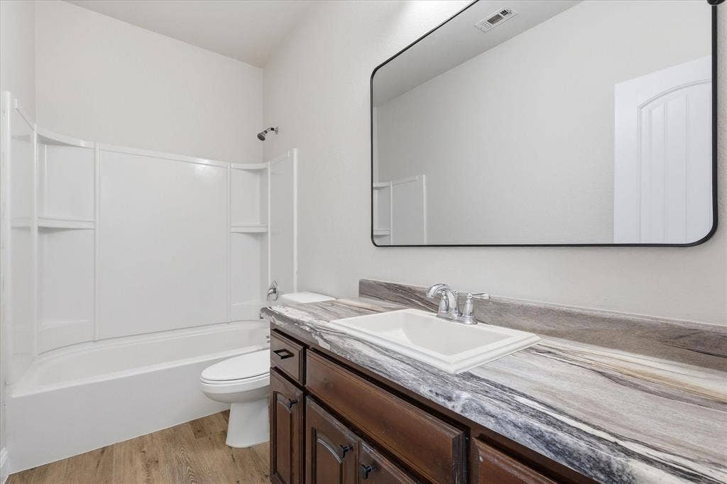 4516 Remuda Lane, Denton, TX 76226 Wood-finish flooring, dark wood vanity with white rectangular sink, framed rectangular mirror, and a white tub with shower