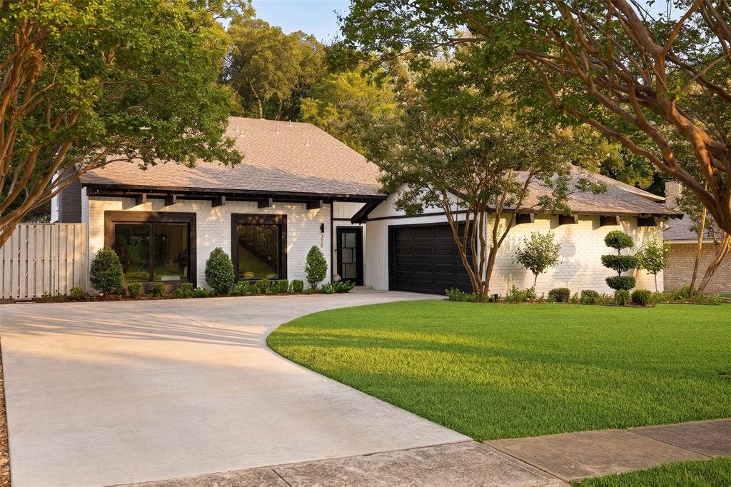 White brick exterior with dark trim accents, curved concrete driveway, attached garage with dark door, green lawn, and manicured landscaping