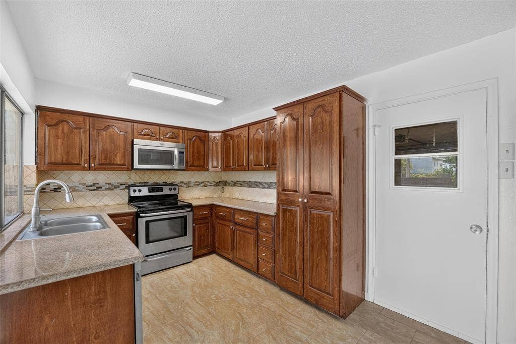 2130 Bowie Drive, Carrollton, TX 75006 Kitchen featuring wood cabinetry, stainless steel appliances, patterned tile backsplash, double basin sink, and light-toned flooring