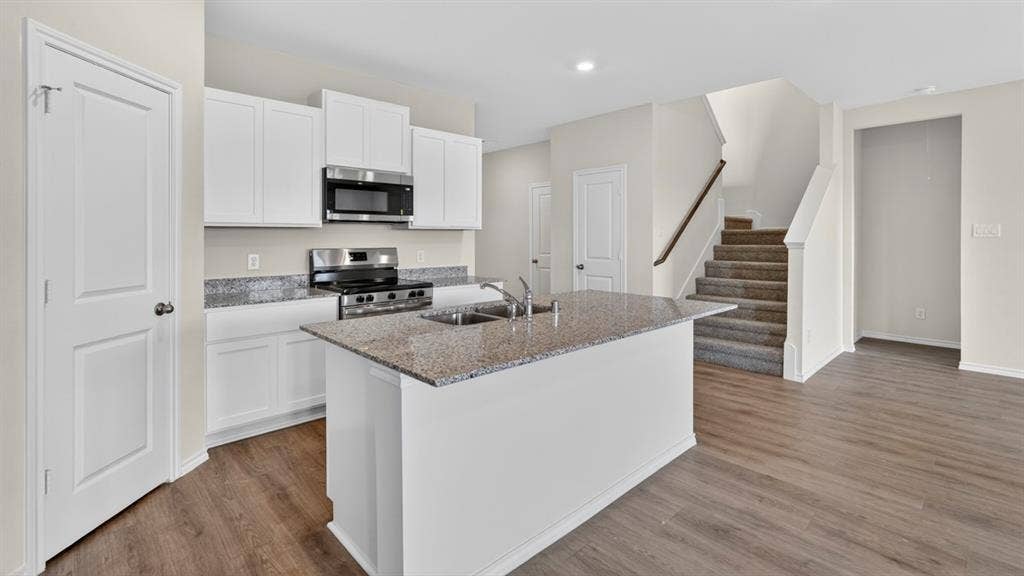 237 Yates Street, Bullard, TX 75757 Modern kitchen featuring white cabinetry, stainless steel appliances, a center island with a sink, wood-finish flooring, and a carpeted staircase