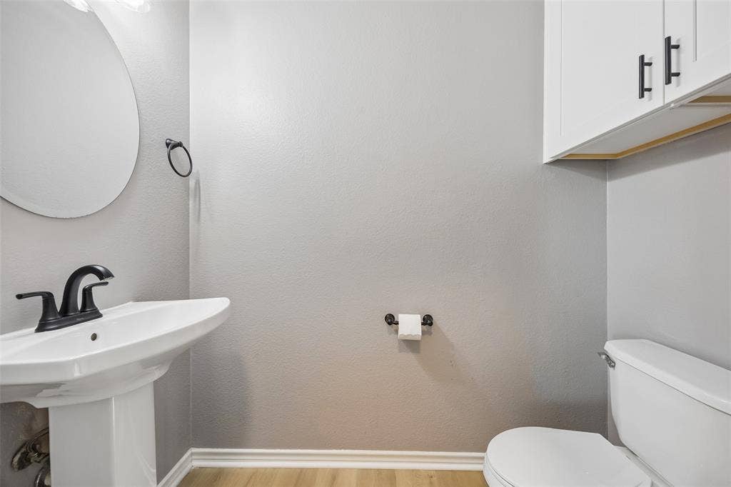 753 Summerfield Drive, Allen, TX 75002 Pedestal sink with a matte black faucet, wall-mounted circular mirror, white cabinetry with black hardware, and wood-finish flooring