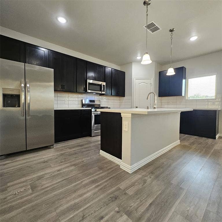 2225 Crooked Bow Drive, Mesquite, TX 75149 Kitchen with wood-finish flooring, dark cabinetry, stainless steel appliances, a white subway tile backsplash, and an island with a built-in sink