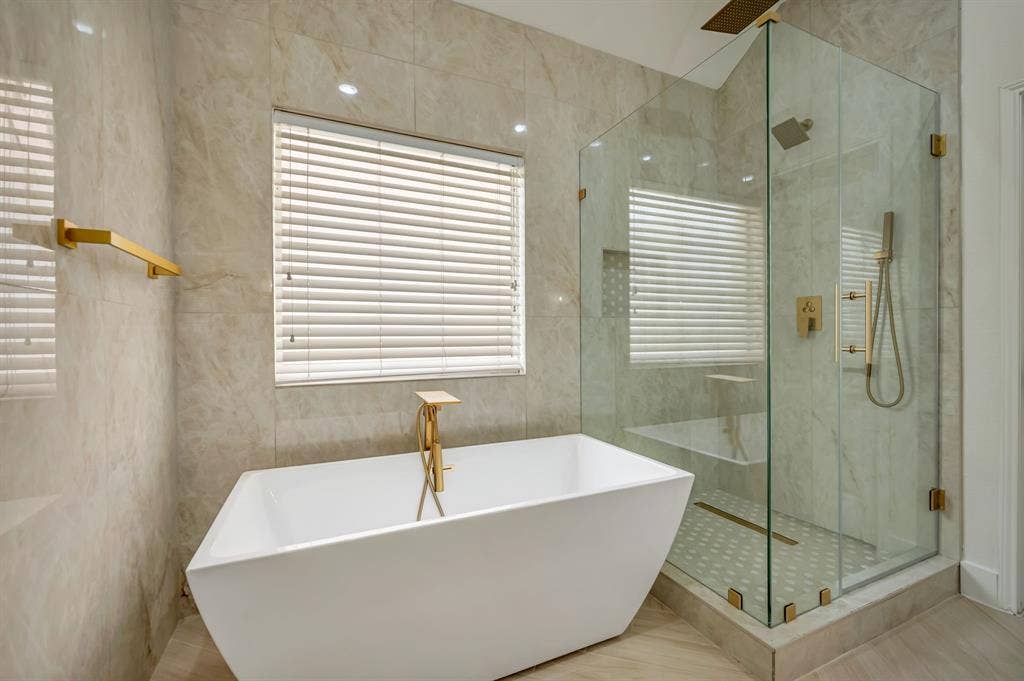 7904 Blacktail Trail, McKinney, TX 75070 Freestanding soaking tub with gold-finish fixtures, a spacious glass-enclosed shower with a built-in bench, and floor-to-ceiling tile surround