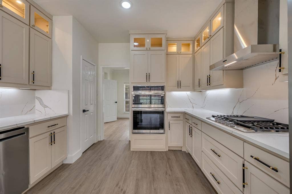 7904 Blacktail Trail, McKinney, TX 75070 Gourmet kitchen featuring a built-in double oven, gas cooktop, stainless steel range hood, stone countertops with matching backsplash, and custom cabinetry with glass-front uppers