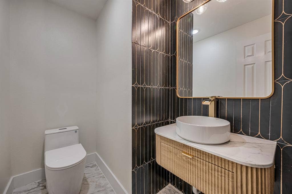 7904 Blacktail Trail, McKinney, TX 75070 Powder room featuring a sleek toilet, patterned wall tile, gold-framed mirror, floating vanity with ribbed Luxury Vinyl Plank cabinetry, and a countertop vessel sink with a gold-tone faucet