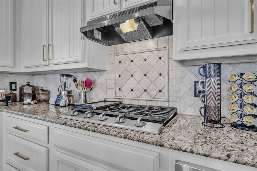 White shaker cabinetry with gold-tone hardware, stainless steel range hood, stone-finish countertops, tiled backsplash with decorative inset, and a built-in gas range