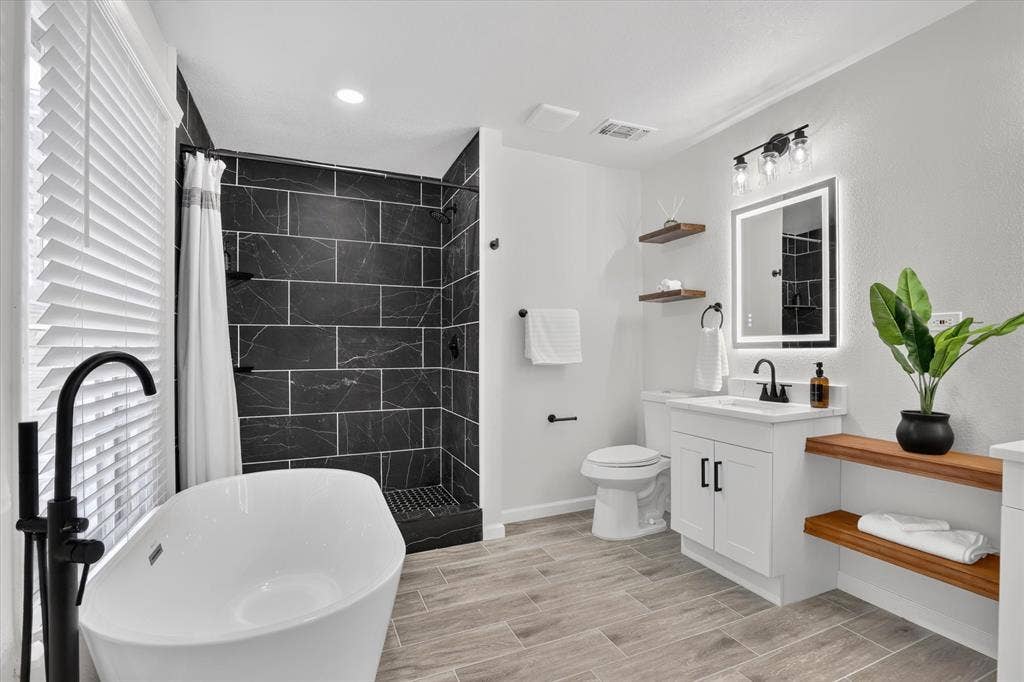 1204 Loma Alta Place, Cleburne, TX 76033 Freestanding soaking tub with a floor-mounted matte black faucet, walk-in shower with dark subway tile, single vanity with a white countertop, and wood-finish tile flooring