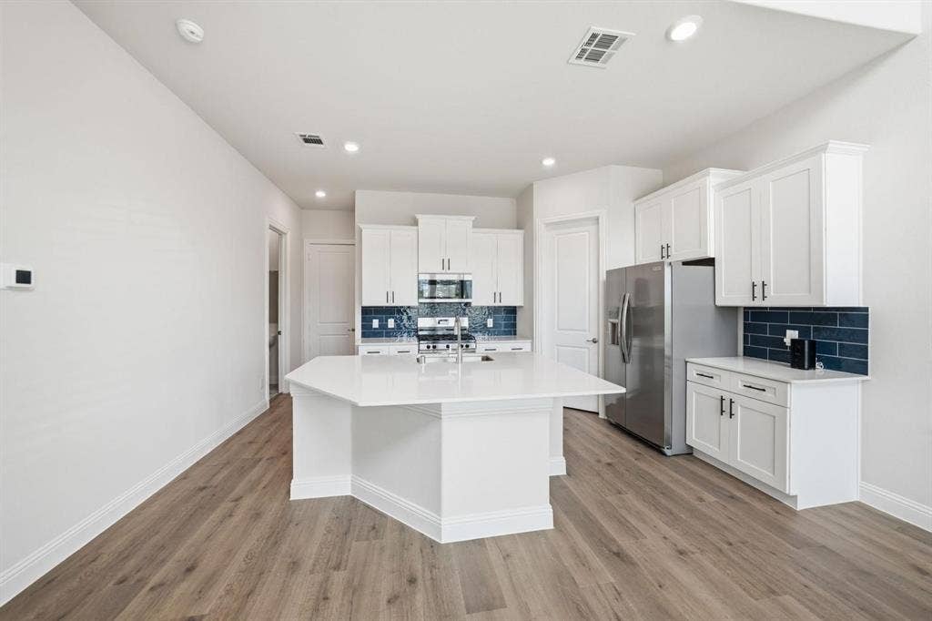 4532 Community Court, Plano, TX 75024 Modern kitchen featuring an angled island with a white countertop, white cabinetry with dark hardware, stainless steel appliances, and a blue subway tile backsplash