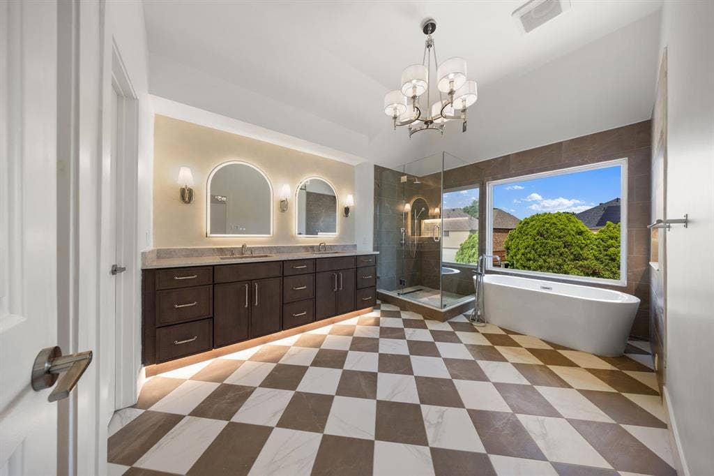 7504 Hughes Drive, Plano, TX 75024 Spacious bathroom featuring checkered tile flooring, a freestanding soaking tub, a frameless glass shower enclosure, a dual vanity with undermount sinks, and illuminated arched mirrors