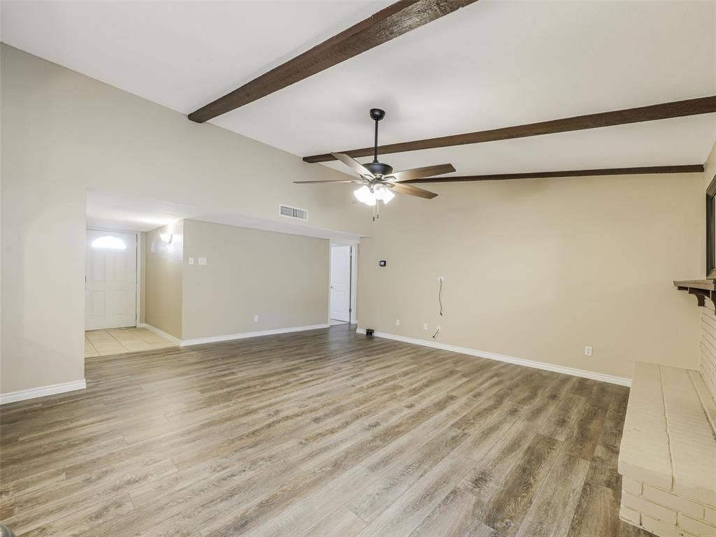 Vaulted ceiling with exposed wood beams, wood-finish flooring, ceiling fan with integrated lighting, brick fireplace with mantel, and recessed entry