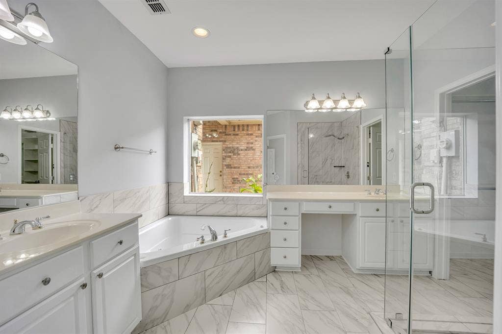 Spacious bathroom featuring a built-in corner tub, glass-enclosed shower, dual vanities with white cabinetry, and large format tile flooring