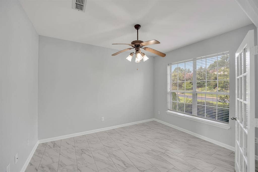 Wood-finish ceiling fan with integrated lighting, large window with blinds, white trim, light gray walls, and light-toned tile flooring