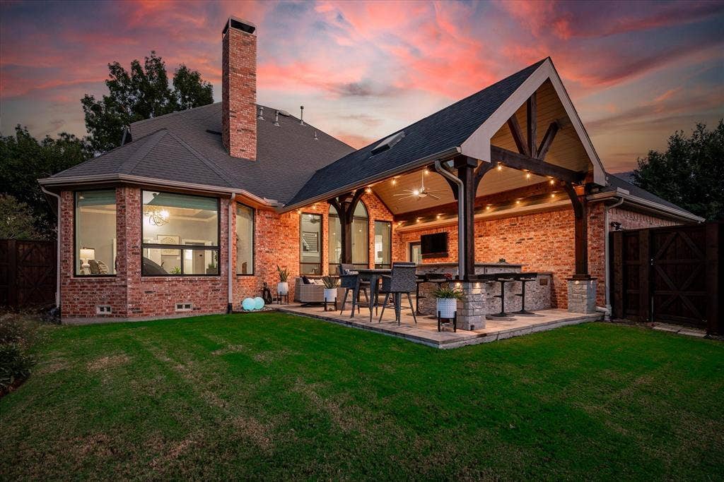 7525 Cedar Elm Drive, Irving, TX 75063 Brick exterior with a prominent chimney, large bay windows, and a covered patio featuring exposed wood beams, a built-in outdoor kitchen, and string lighting