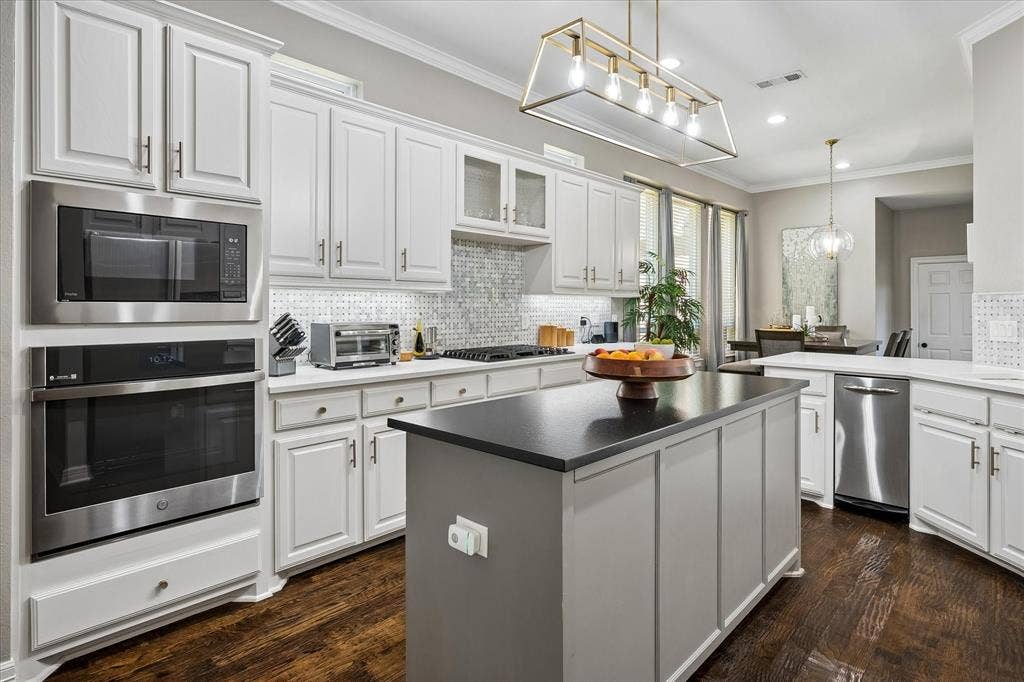 7525 Cedar Elm Drive, Irving, TX 75063 White cabinetry with brushed nickel hardware, stainless steel appliances, dark wood-finish flooring, a kitchen island with a dark countertop, and a tiled backsplash