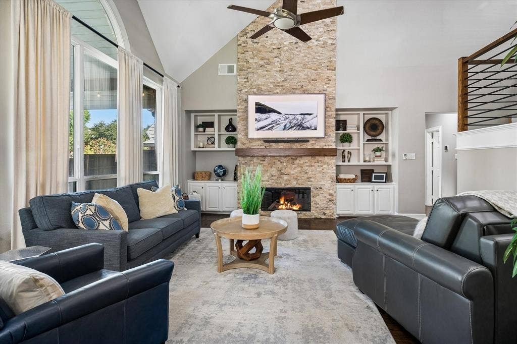 7525 Cedar Elm Drive, Irving, TX 75063 Vaulted ceiling with a ceiling fan, stone-clad fireplace with a wood mantle, built-in shelving and cabinetry, and large arched windows