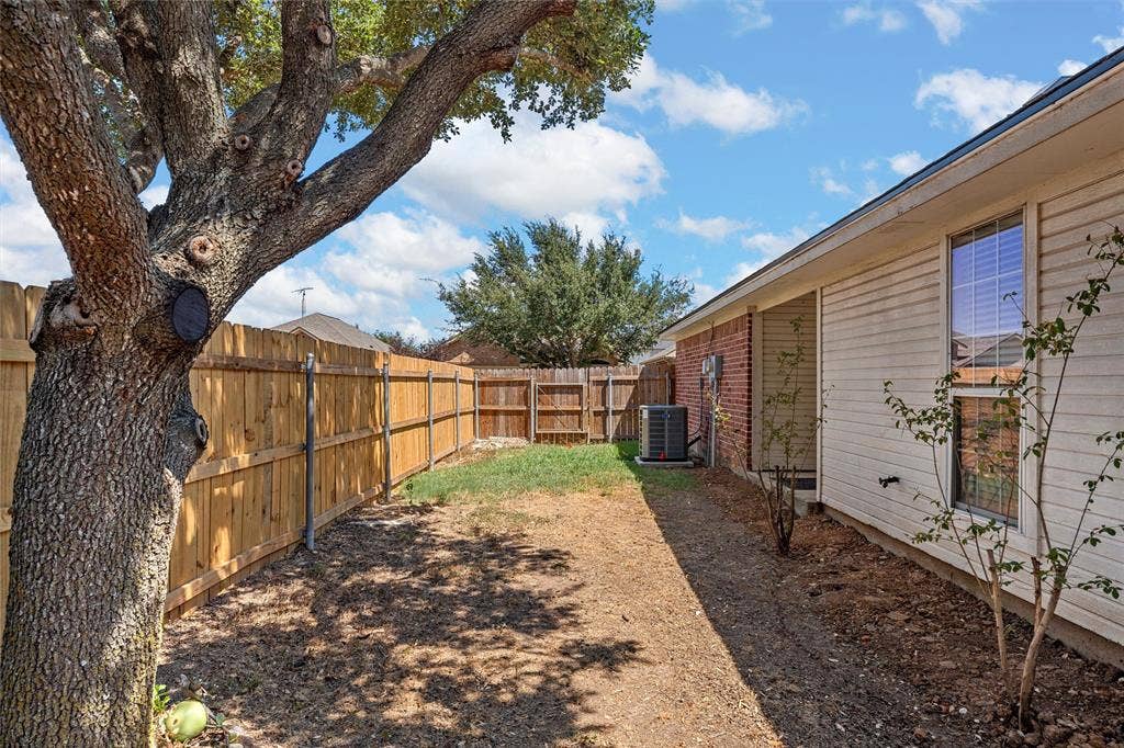 Fully fenced backyard featuring a large mature tree, established shrubbery, and brick and siding exterior