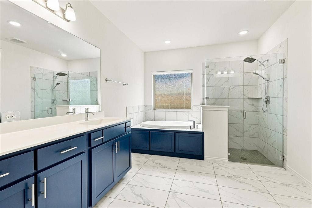 2812 Dunbar Drive, Northlake, TX 76247 Dual vanity with blue cabinetry and white countertops, enclosed glass shower with tiled walls and rain showerhead, soaking tub with tiled surround, and white tile flooring