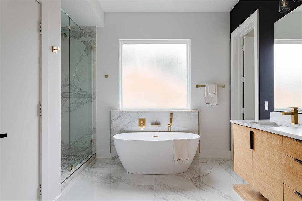 939 N Buckner Boulevard, Dallas, TX 75218 Freestanding soaking tub with wall-mounted brass fixtures, glass-enclosed shower with marble-look tile, double vanity with light wood-finish cabinetry, white countertop, and brass faucets, and marble-look floor tile throughout